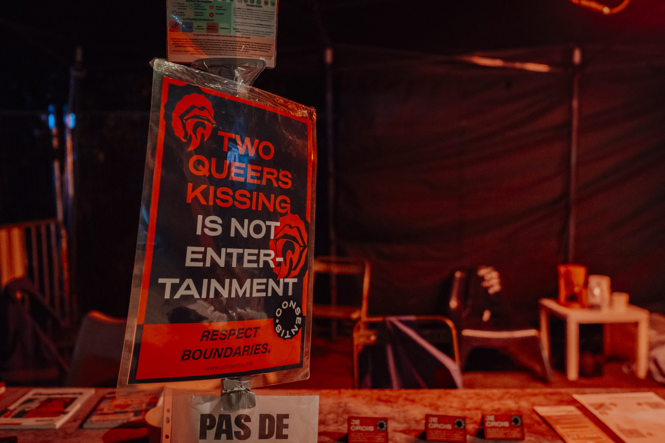 Photo de nuit d’un stand éclairé en rouge. Au centre, une affiche plastifiée sur un support affiche « TWO QUEERS KISSING IS NOT ENTERTAINMENT » et « RESPECT BOUNDARIES », avec le logo Consentis. En bas, on voit une feuille avec le texte « PAS DE … ». Une table avec des documents est floue au premier plan.