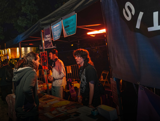 Scène de nuit sous une tente de stand. Deux personnes discutent face à face au-dessus d’une table couverte de flyers et d’objets. Des affiches et pancartes sont accrochées au bord de la tente. À droite, une grande banderole sombre affiche le mot « CONSENTIS » partiellement visible. Éclairage orange.