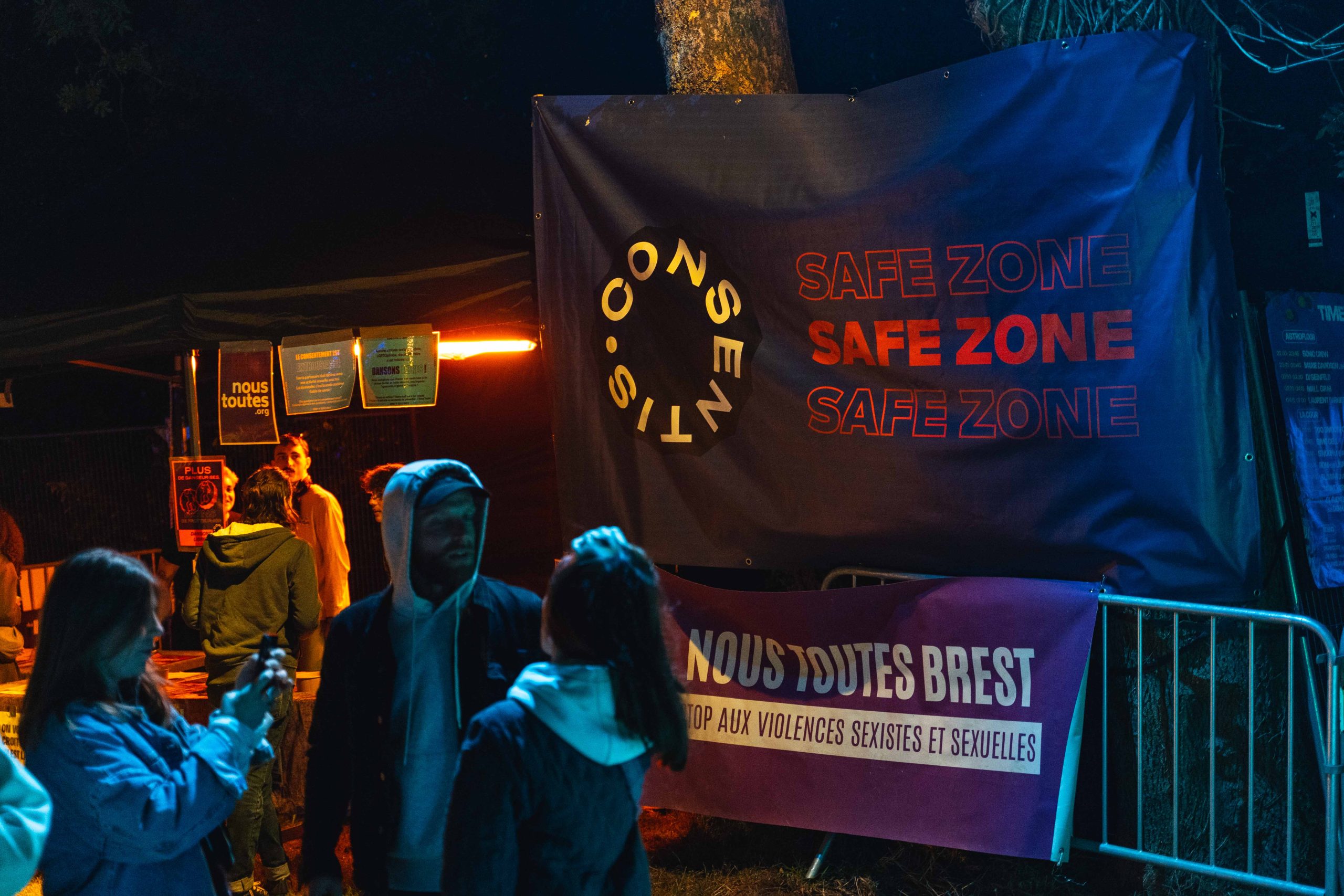 Photo de nuit d’un stand en extérieur. Une grande banderole bleu foncé affiche « SAFE ZONE » trois fois et le logo « CONSENTIS ». En dessous, une banderole violette indique « NOUS TOUTES BREST » et « STOP AUX VIOLENCES SEXISTES ET SEXUELLES ». Plusieurs personnes se tiennent devant des barrières, avec un éclairage bleu et orange.