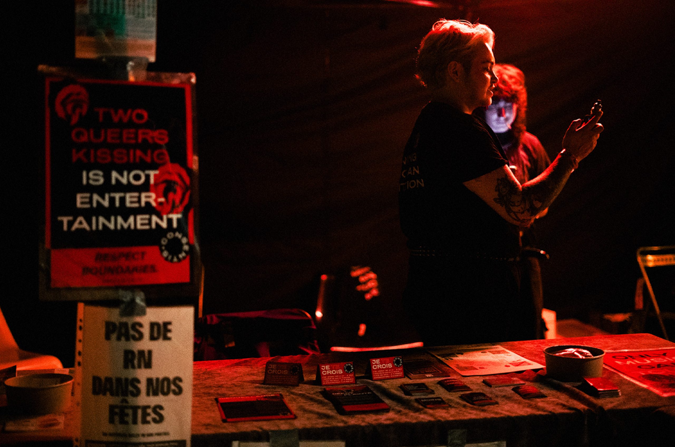 Photo de nuit d’un stand avec éclairage rouge. Une table recouverte d’une nappe présente des flyers et des cartes. À gauche, un panneau affiche « TWO QUEERS KISSING IS NOT ENTERTAINMENT » et en dessous « PAS DE RN DANS NOS FÊTES ». À droite, une personne debout tient un téléphone, avec une autre silhouette derrière.