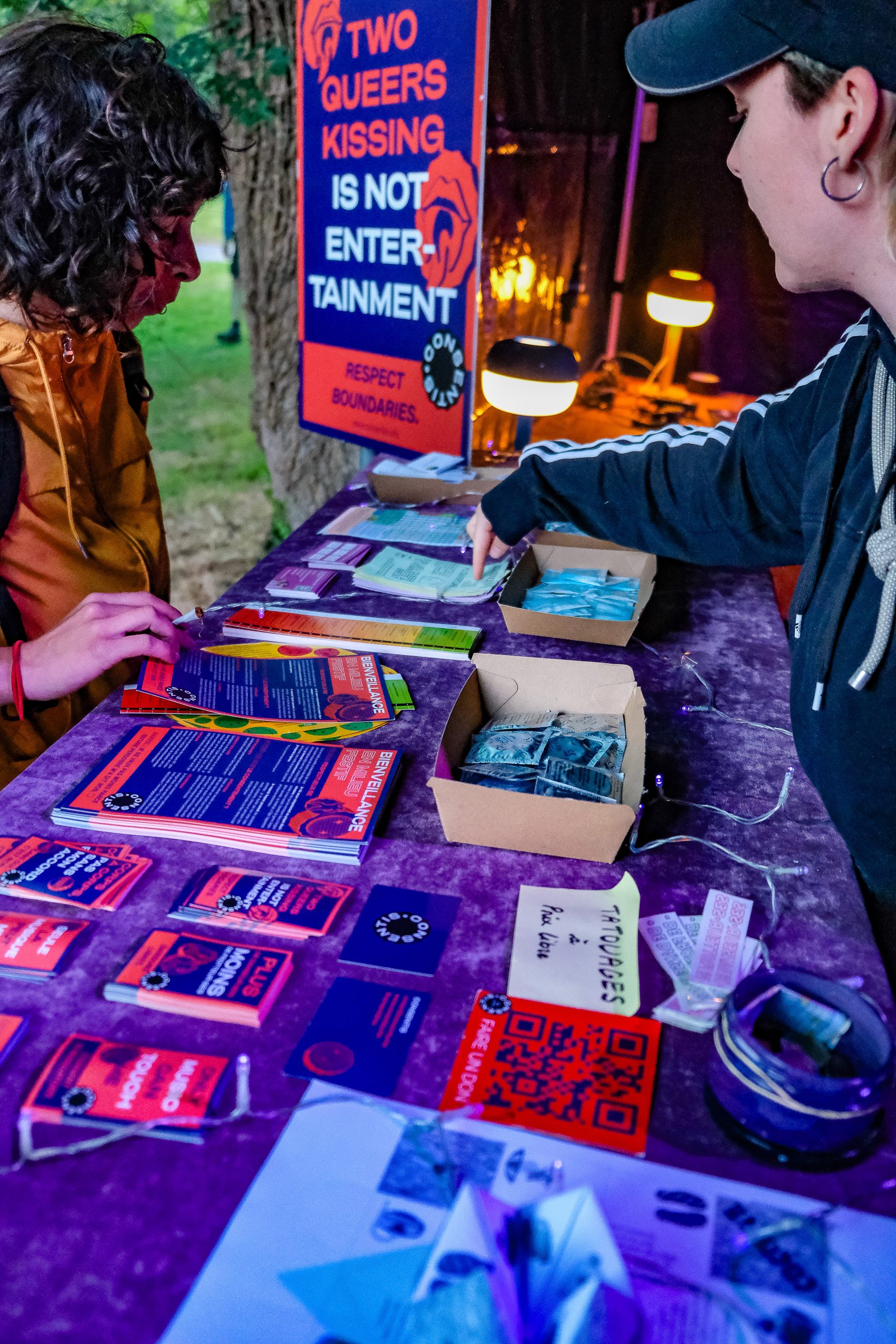 Photo de nuit d’une table de stand recouverte d’une nappe violette, avec des piles de flyers violets et rouges, des cartes, des QR codes et deux boîtes remplies de petits sachets. Deux personnes sont de chaque côté de la table ; l’une pointe du doigt des documents. En arrière-plan, une affiche indique « TWO QUEERS KISSING IS NOT ENTERTAINMENT ».
