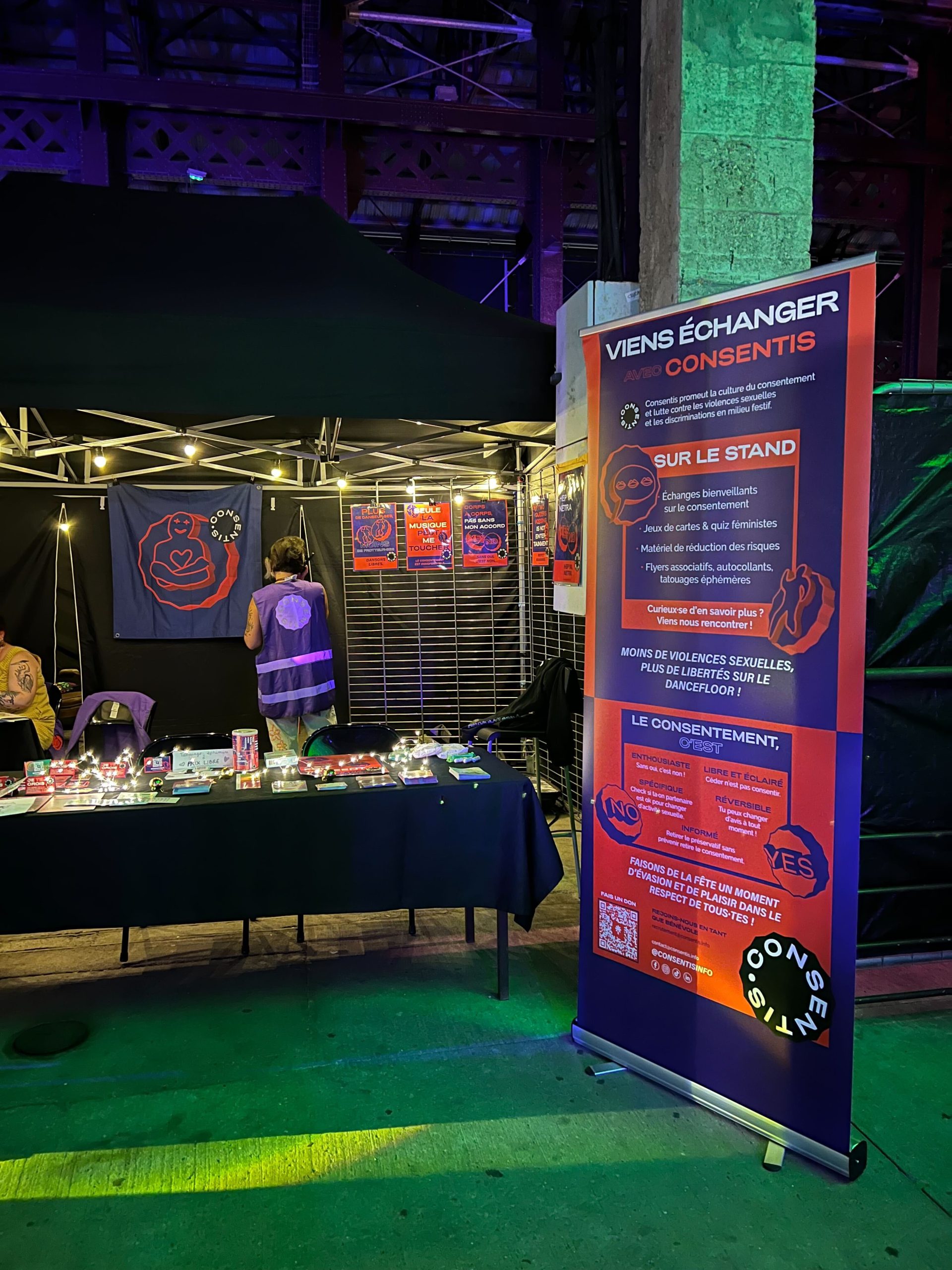 Photo de nuit d’un stand sous une tente noire. À droite, un kakemono violet et orange affiche « VIENS ÉCHANGER AVEC CONSENTIS » avec du texte et des pictogrammes. À gauche, une table recouverte d’une nappe noire présente des flyers et du matériel. Au fond, une banderole et plusieurs affiches sont accrochées sur une grille.