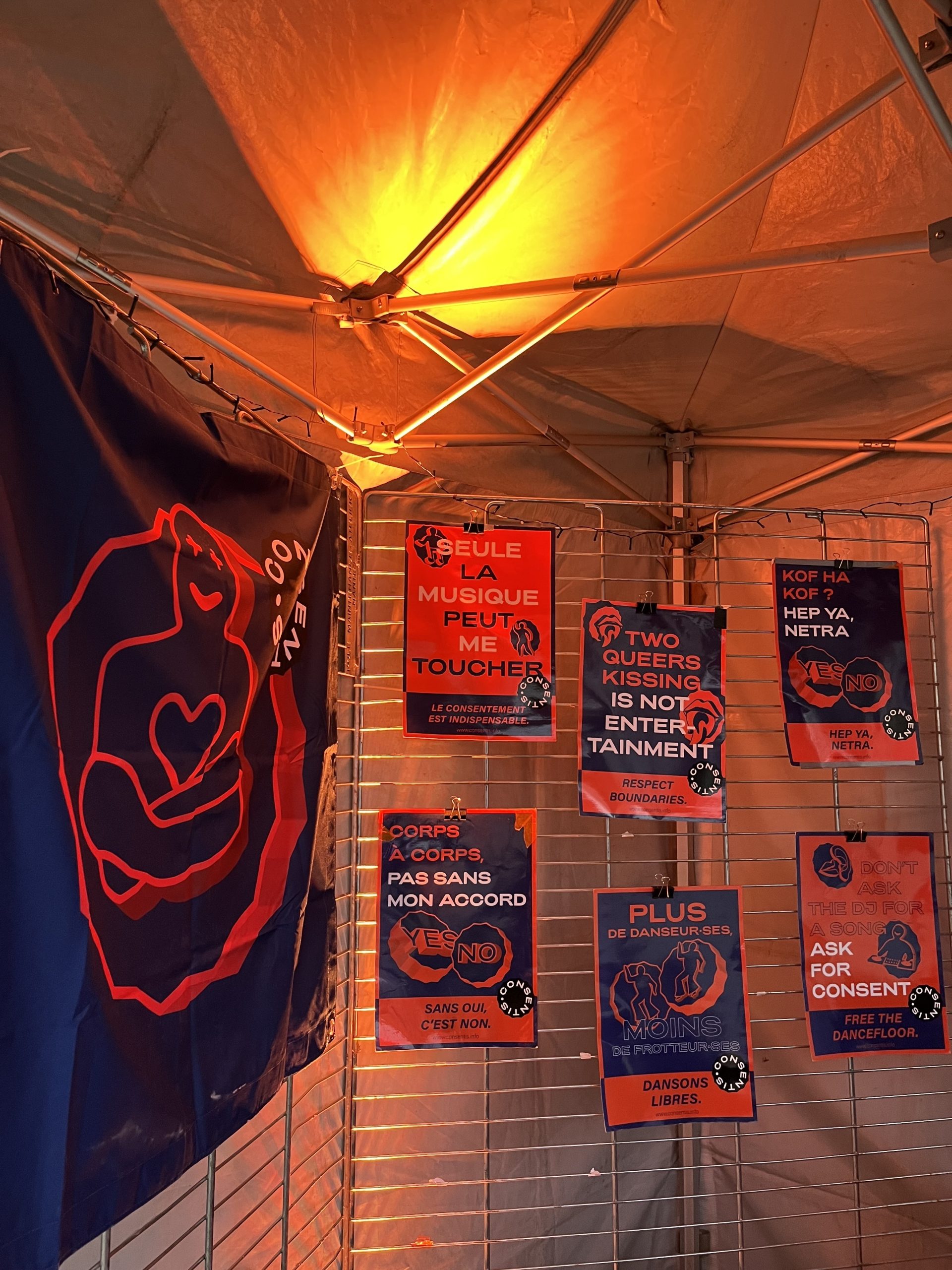 Intérieur d’une tente éclairée en orange. À gauche, une grande banderole bleu foncé et rouge avec un dessin stylisé et le nom « Consentis » partiellement visible. À droite, plusieurs affiches bleu et rouge sont accrochées sur une grille métallique, avec des messages sur le consentement dont « TWO QUEERS KISSING IS NOT ENTERTAINMENT ».