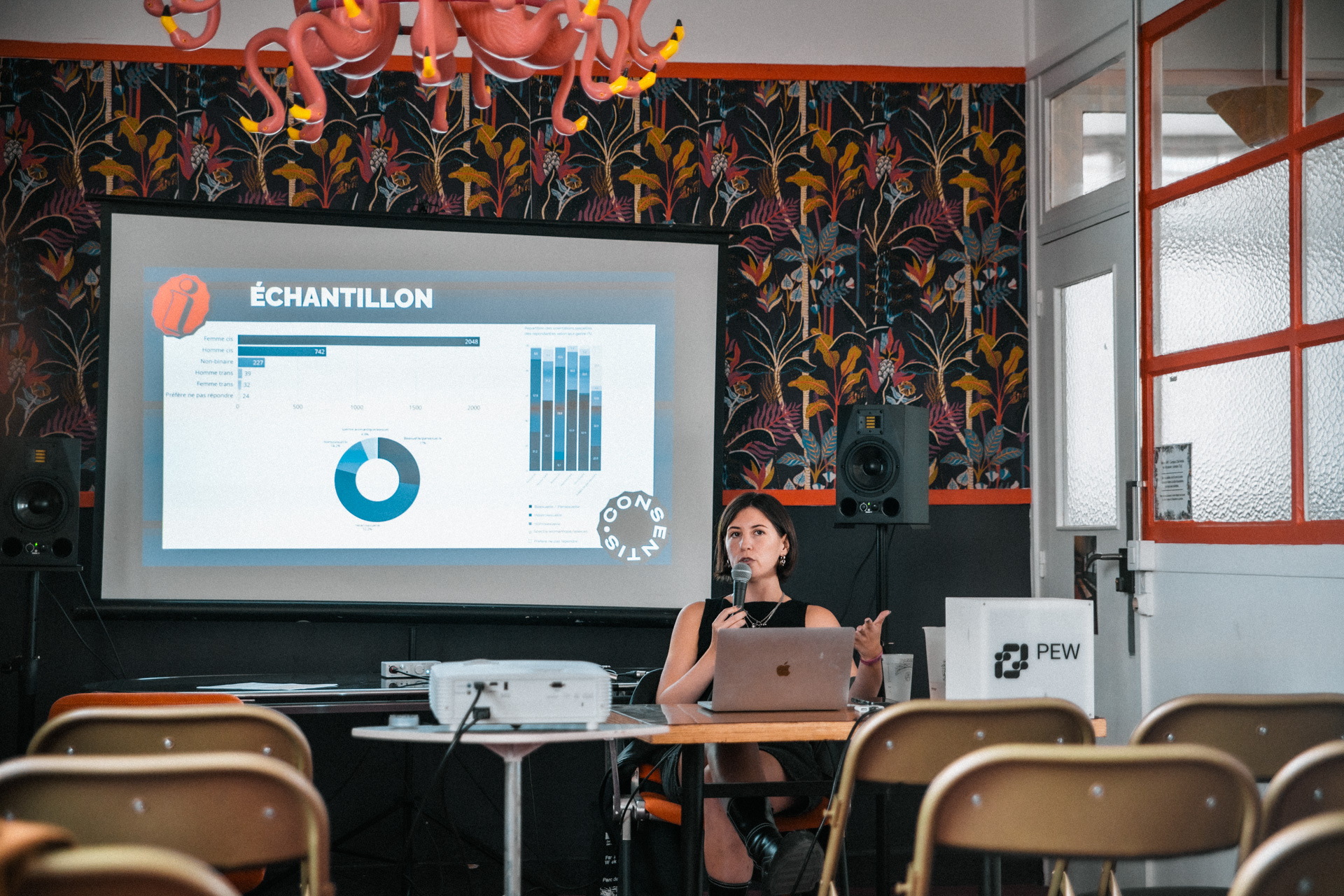 Personne assise derrière une table, parlant dans un micro, avec un ordinateur portable devant elle. Derrière, un écran de projection affiche une diapositive intitulée « ÉCHANTILLON » avec des graphiques. La salle a un papier peint à motifs végétaux, des enceintes de chaque côté et des rangées de chaises au premier plan.