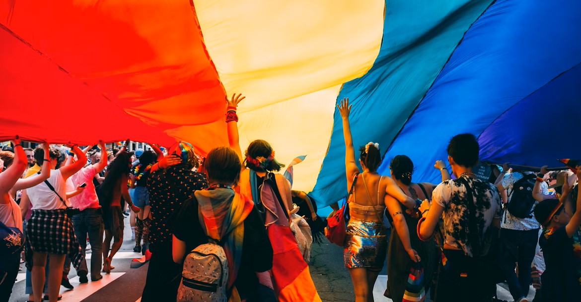 Foule participant à une marche des fiertés sous un grand drapeau arc-en-ciel multicolore, en pleine rue.