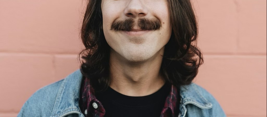 Portrait cadré en gros plan d’une personne souriante à moustache, aux cheveux longs, portant une veste en jean devant un mur rose.