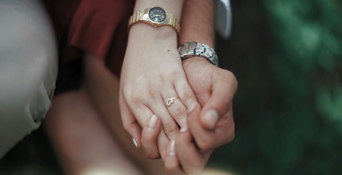 Gros plan sur deux mains entrelacées, avec montres au poignet et bague fine, dans une ambiance douce en extérieur.