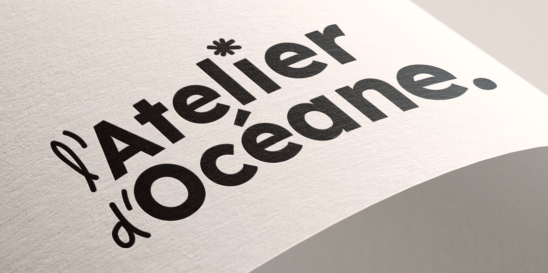 Gros plan d’un papier blanc texturé posé en diagonale, avec le texte « l’Atelier d’Océane » imprimé en noir. Fond gris clair.