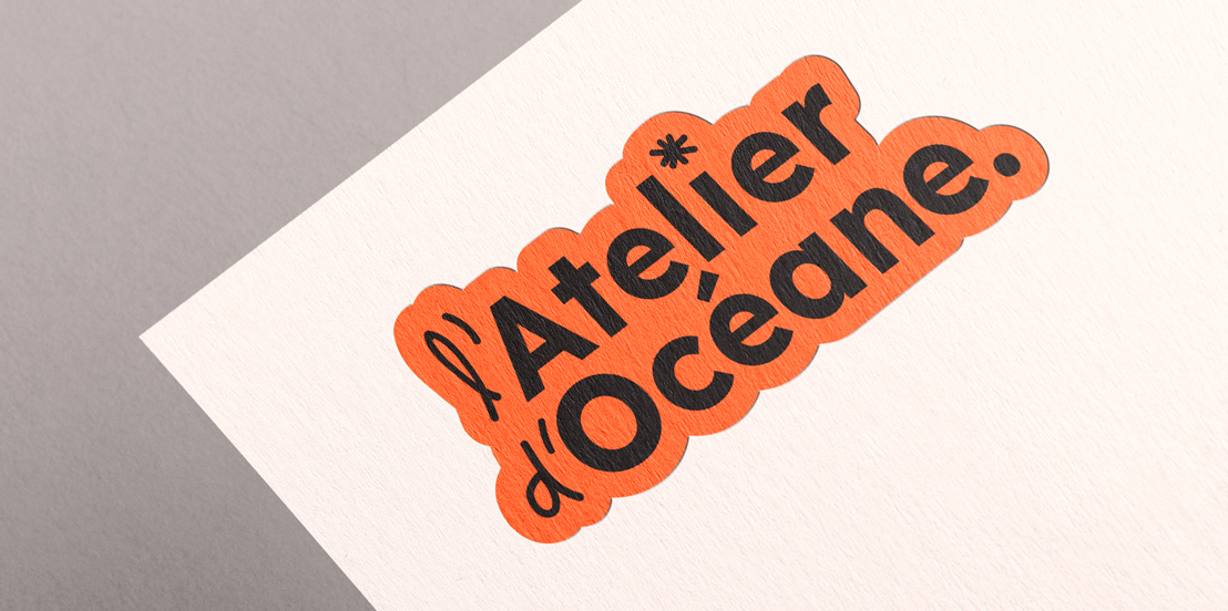Photo en plongée d’une feuille de papier blanc cassé, légèrement granuleuse, disposée en diagonale sur un fond gris. En haut de la feuille, un autocollant orange à bords arrondis affiche le logo « l’Atelier d’Océane. » en lettres noires.