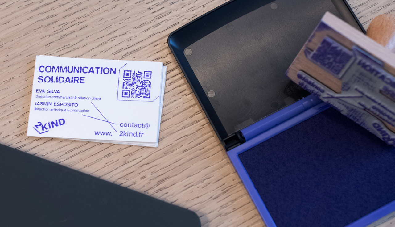 Sur une table en bois clair, on voit une petite pile de cartes de visite blanches imprimées en bleu/violet avec le titre “COMMUNICATION SOLIDAIRE”, un QR code et les noms “Eva Silva” et “Jasmin Esposito”, ainsi que l’adresse “contact@2kind.fr ”. À droite, un tampon encreur ouvert montre une mousse d’encre bleue, avec un tampon en bois partiellement visible au-dessus. En arrière-plan, le coin d’un ordinateur portable noir apparaît, légèrement flou, ce qui met l’accent sur les cartes et le matériel de marquage.