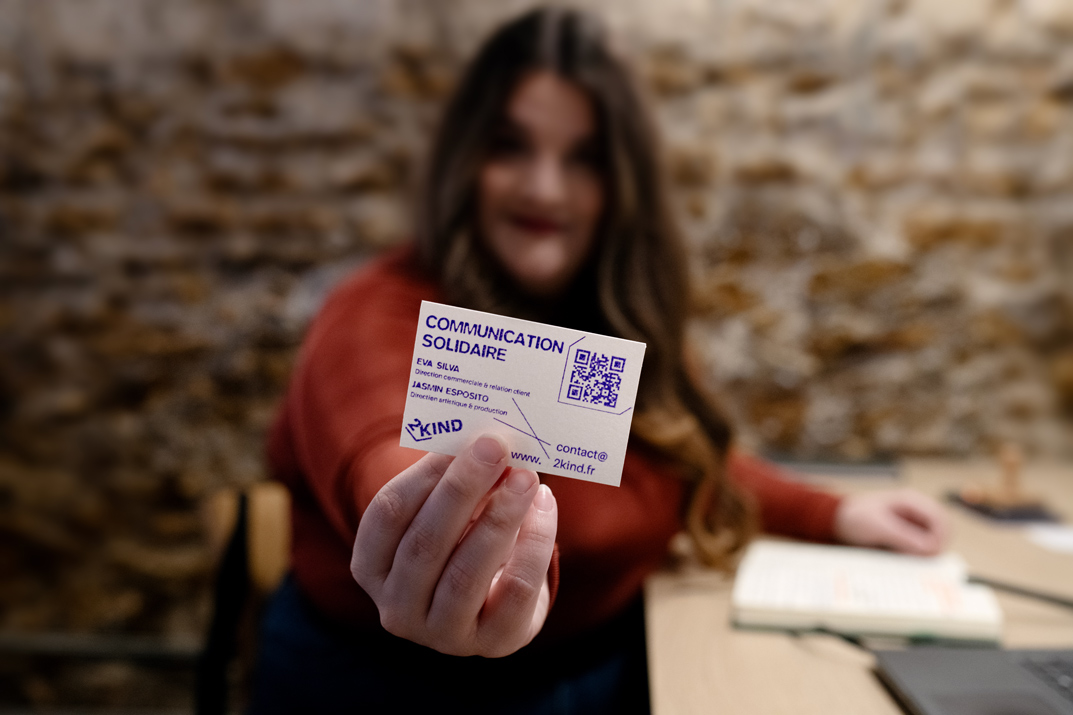 Au premier plan, une main tient une carte de visite blanche et violette de 2KIND, avec le titre « Communication solidaire », les prénoms Eva Silva et Jasmin Esposito, un QR code et l’adresse de contact. L’appareil fait la mise au point sur la carte, qui remplit le centre de l’image. À l’arrière-plan, volontairement flou, on distingue une femme en haut rouge assise à une table, un carnet ouvert et un ordinateur, devant un mur en pierres apparentes.