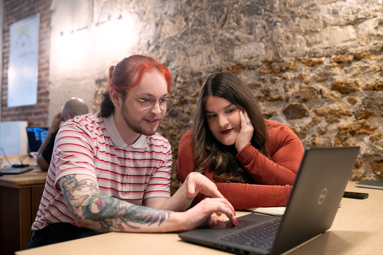Deux personnes assises à une table en bois regardent un ordinateur portable ouvert. La personne à gauche, avec des cheveux rouges attachés et un polo blanc à rayures roses, pointe l’écran; la personne à droite, aux cheveux longs bruns et en haut rouge brique, appuie sa joue sur sa main. Mur en pierre derrière.
