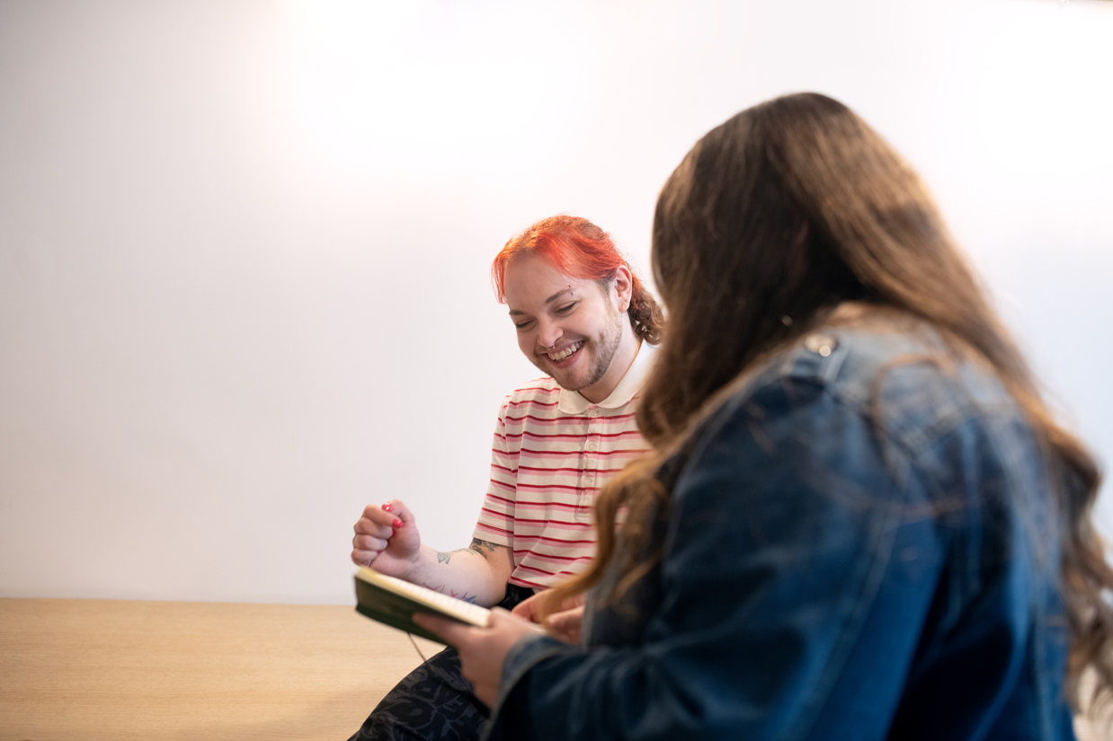 Photo prise en intérieur, sur fond de mur blanc et d’une grande table en bois. Une personne aux cheveux roux, assise, sourit et semble échanger de façon conviviale. Au premier plan (flou), une autre personne en veste en jean tient un carnet ou agenda ouvert, comme si elle prenait des notes ou présentait un point. L’ambiance évoque un moment de travail ou de brainstorming.