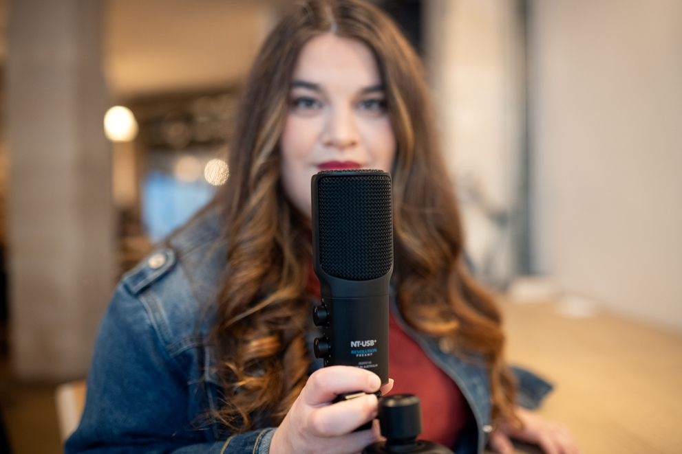 Portrait en intérieur : le micro noir (type micro USB) est au premier plan et net, tenu à bout de bras par une femme aux cheveux longs ondulés. Son visage apparaît légèrement flou en arrière-plan, avec une expression calme. Le décor ressemble à un bureau/coworking, éclairé doucement, avec des éléments de fond volontairement floutés (profondeur de champ).