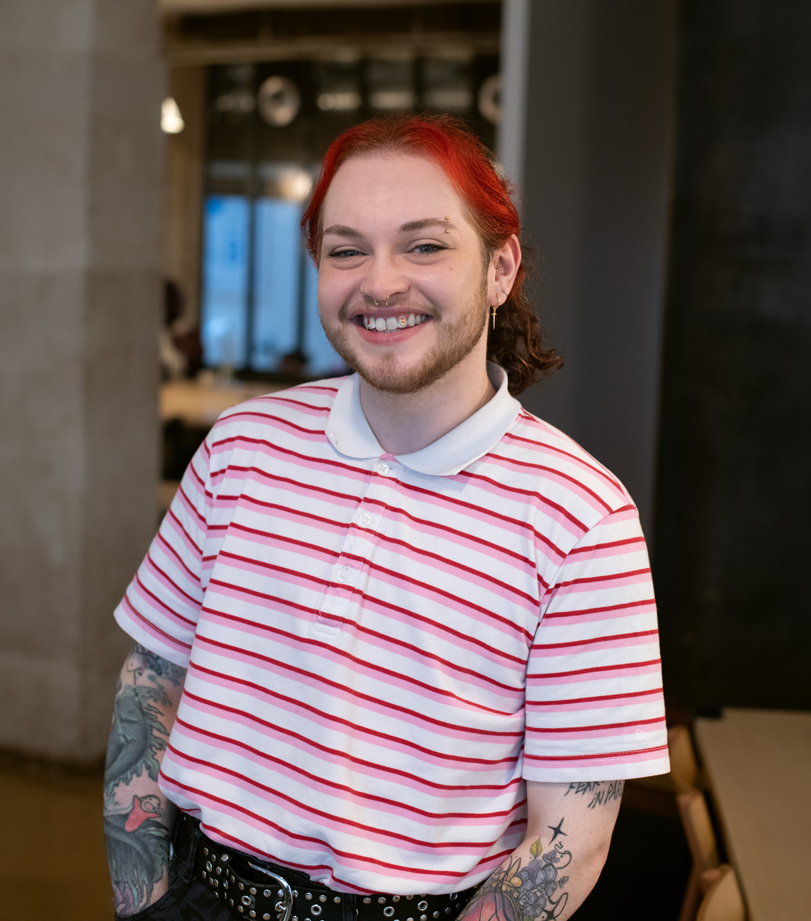 Portrait d’une personne souriante aux cheveux rouges, portant un polo blanc à rayures roses, un pantalon sombre et une ceinture noire à studs, avec des tatouages visibles sur les avant-bras, devant un intérieur de bureau flou.