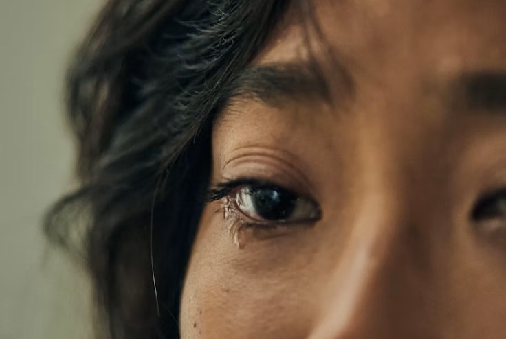 La photo montre un cadrage très serré sur une partie du visage, centré sur un œil humide. Une larme brillante descend sous l’œil, tandis que les cils et la peau autour sont nets et détaillés. L’arrière-plan est flou et la lumière douce renforce l’émotion de tristesse.