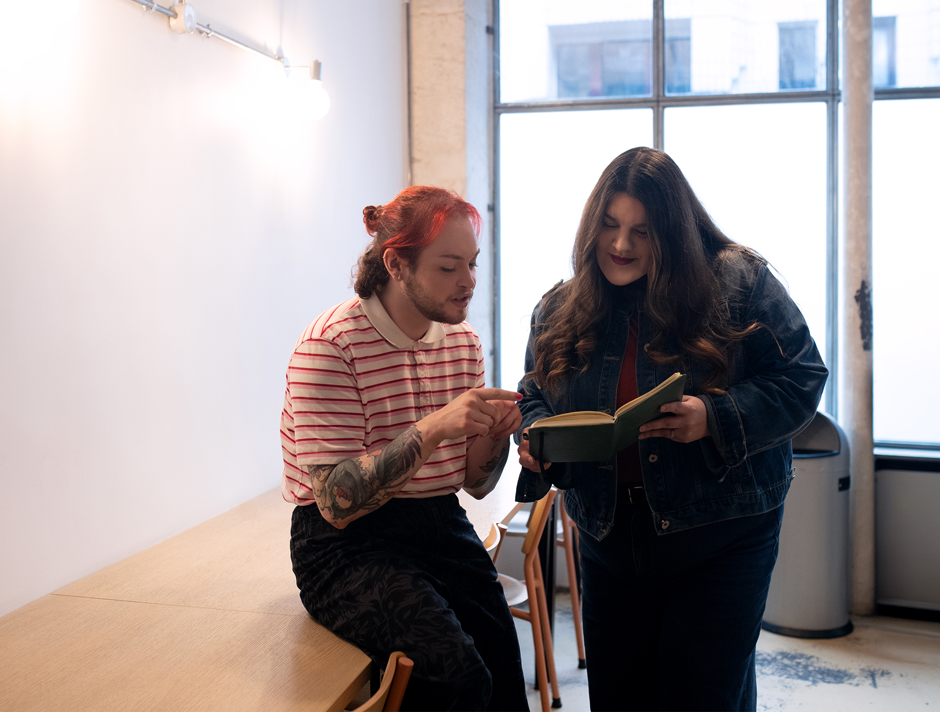 Deux personnes près d’une grande fenêtre dans une pièce claire ; l’une est assise sur une table en bois et pointe du doigt un carnet ouvert, l’autre est debout en tenant le carnet, toutes deux regardant les notes.