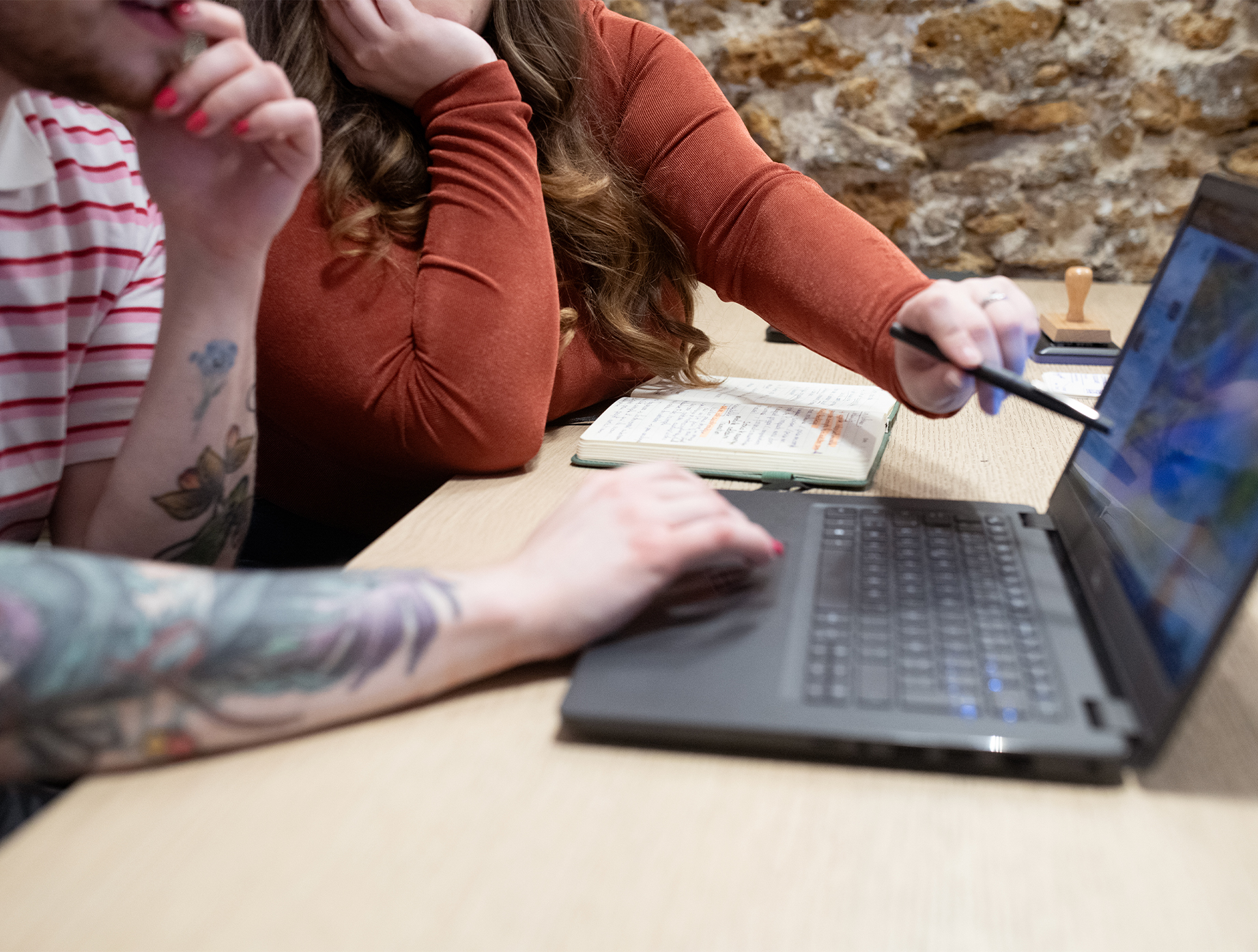 Gros plan sur une table en bois : un ordinateur portable ouvert au premier plan, une main tatouée posée près du clavier, et une autre main tenant un stylet pointe l’écran. Un carnet ouvert et un tampon en bois sont posés derrière, devant un mur en pierres.