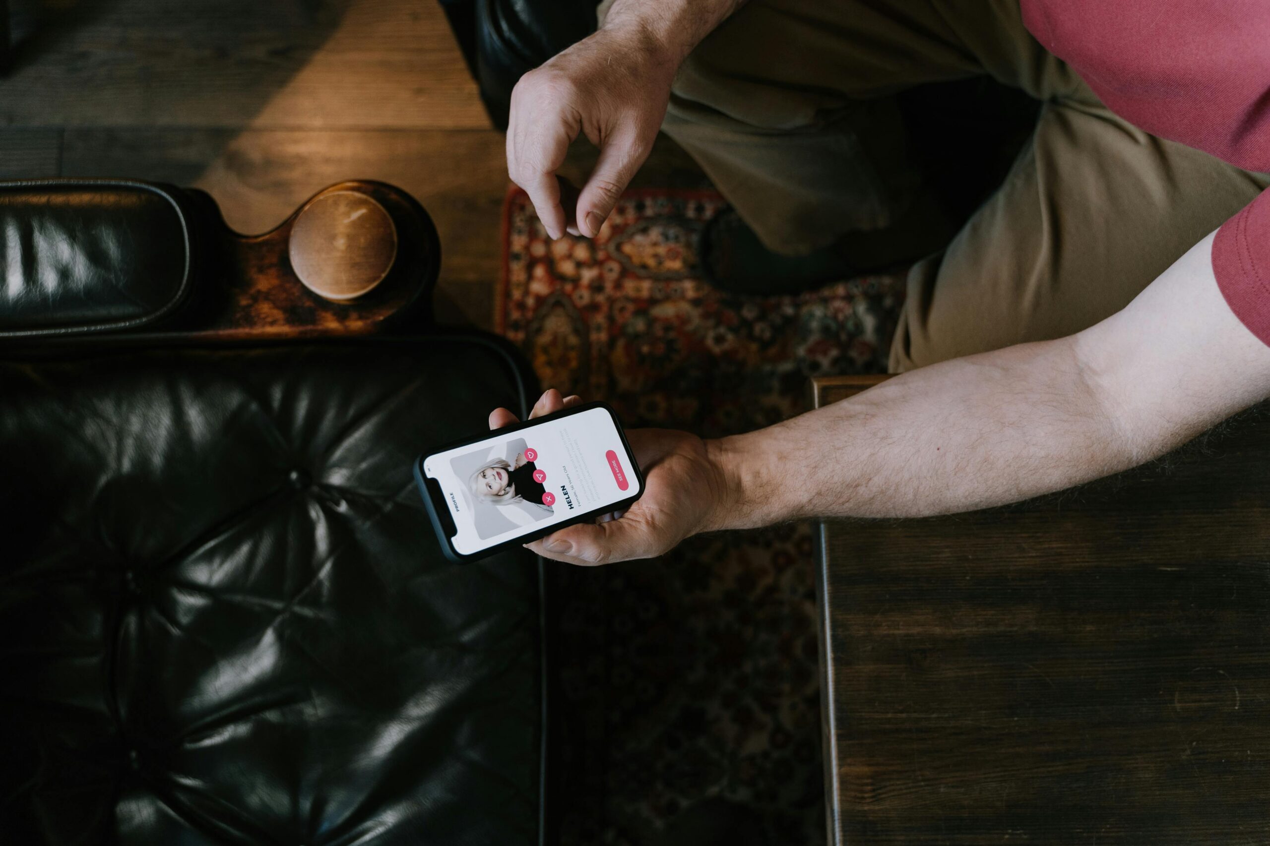 Vue du dessus d’une personne assise, tenant un smartphone affichant une application, près d’un canapé en cuir noir.